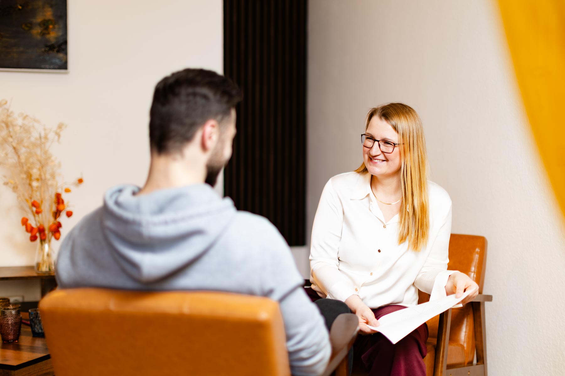 Eine Frau mit langen blonden Haaren sitzt einem jungen Mann gegenüber und erklärt ihm etwas anhand eines Zettels. Sie lächelt freundlich.